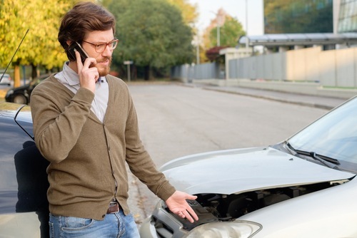 Young man on his cell phone with his insurer after a car wreck. He is standing between the two involved cars, one of which has a buckled front hood
