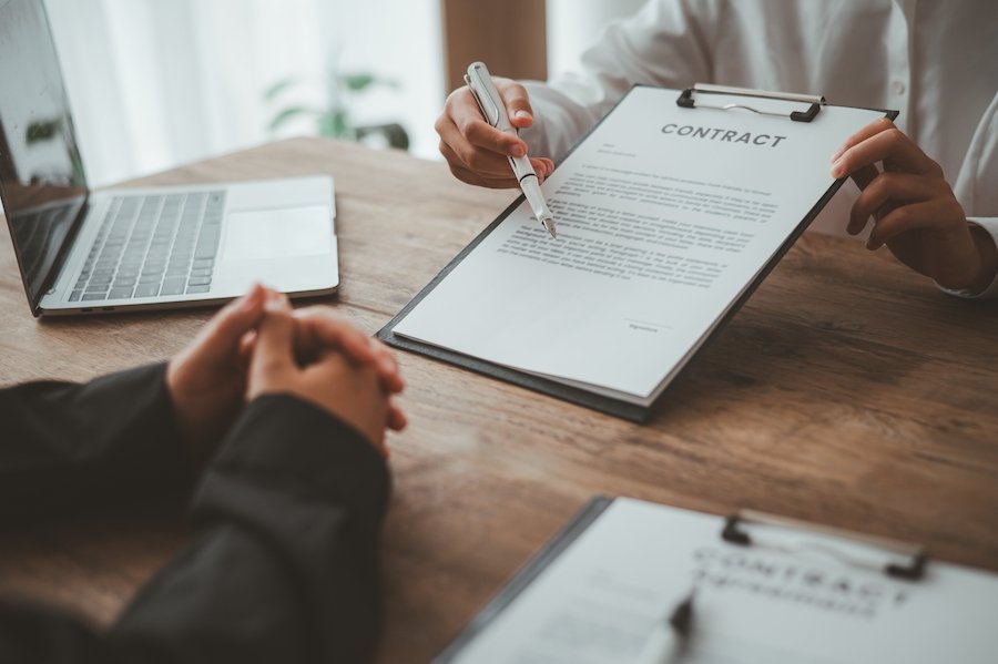 Closeup of hands above a desk holding up a clipboard with a document entitled "contract", and person with crossed hands is looking at it, and there is a laptop in the background