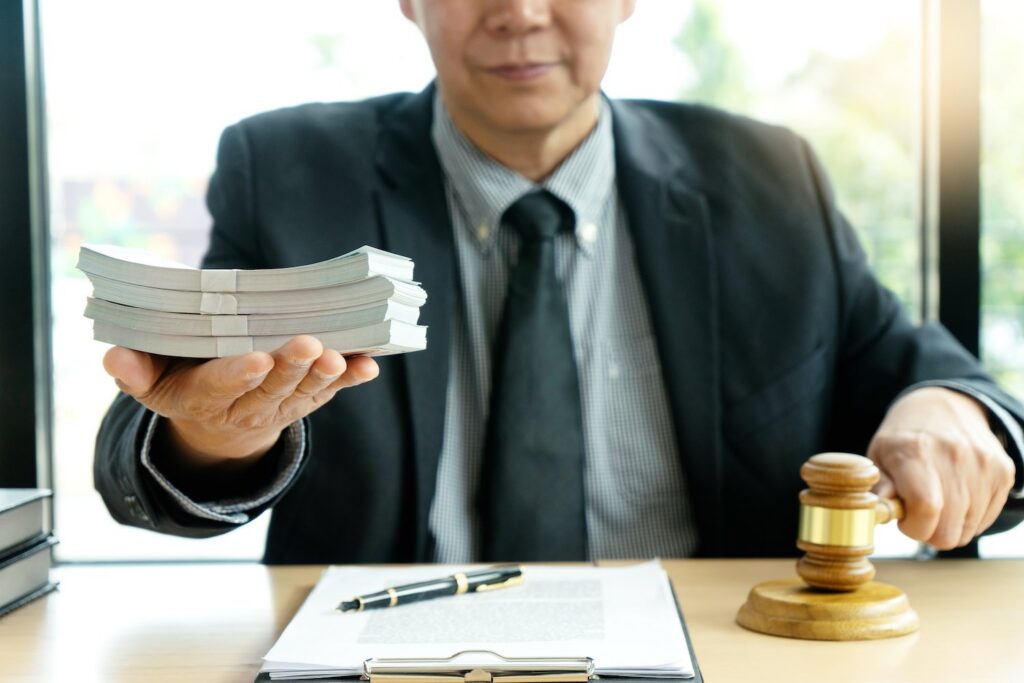 Judge in a business suit seated at a table with a clipboard in front of him, and he is holding a gavel in one hand and a wad of cash in the other, and an office window is behind him